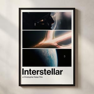 Op de afbeelding: Een ingelijste poster voor de film "Interstellar", met drie horizontale panelen met scènes uit de ruimte. De titel "Interstellar" staat in vette zwarte letters, met de naam van de regisseur eronder. De lijst is zwart.