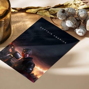 May include: A Captain America print featuring the superhero in a battle scene. The print has the words "CAPTAIN AMERICA" in white text. The image is on a dark background with a fiery glow. A decorative glass bottle and dried floral arrangement are also in the image.