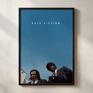以下が含まれることがあります： 鮮やかな青い空を背景に、映画のタイトル「パルプ・フィクション」が描かれた額入りのポスター。黒いスーツを着た2人の男性が下を見ている画像です。ポスターは黒いフレームに入っています。