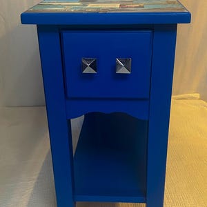 May include: A bright blue side table featuring a single drawer with two silver pyramid-shaped knobs. The top surface is decorated with a collage of various rectangular images. The table includes a lower open shelf and four straight legs.