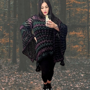 May include: A black, purple, and green crochet poncho with bell sleeves. The poncho features a geometric pattern and is worn with black leggings and platform shoes. The background is a blurred forest scene.