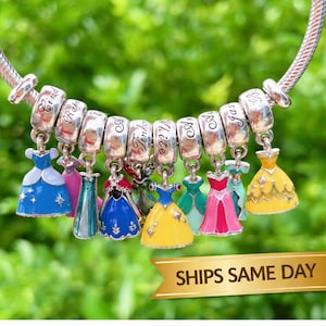 May include: A silver-tone charm bracelet featuring multiple dangling dress charms in various colors including blue, yellow, pink, and green. Each charm is attached to a silver bead engraved with cursive text, showcasing a collection of miniature gowns.