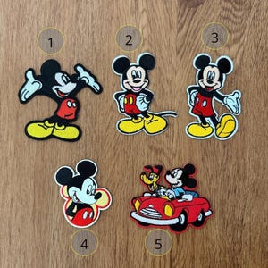 Może przedstawiać: Pięć haftowanych naszywek z Myszka Miki w różnych pozach. Miki ma na sobie klasyczne czerwone szorty, żółte buty i czarne rękawiczki. Jedna naszywka przedstawia Mikiego prowadzącego czerwony samochód z inną postacią.