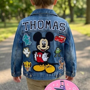 May include: A blue denim jacket featuring the name THOMAS in white letters with black outlines across the top. The back is decorated with embroidered patches of Mickey Mouse, a red race car, a green monster, a white glove, and a lion cub.