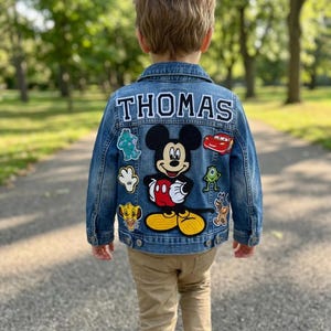 May include: A denim jacket with the name "THOMAS" in white letters on the back. The jacket features patches of Mickey Mouse, a red race car, and other cartoon characters. The jacket is blue and has a classic denim design.