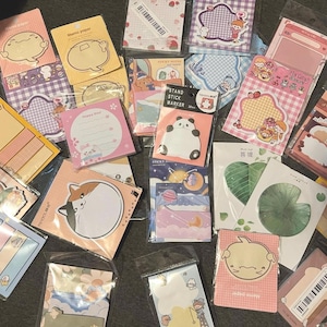 May include: Assortment of colorful sticky note pads with various designs, including cartoon characters, patterns, and shapes. The pads are in different sizes and colors, such as pink, yellow, and blue. Some pads have text, and some have a barcode.