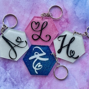 May include: A collection of hexagonal keychains with silver keyrings. Each keychain features a glittery background in white, pink, and blue, with a black or white cursive letter design. The letters are J, L, H, and Z.