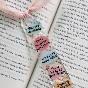 May include: A clear acrylic bookmark with a pink ribbon, featuring motivational quotes in various colors. The quotes include "You are awesome," "Don't be afraid just believe," and "Anything is possible."