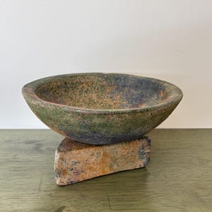 Puede incluir: Un cuenco ovalado rústico con superficie texturizada, en tonos verdes, marrones y azules, apoyado sobre una base de piedra rectangular. La base de piedra muestra tonos anaranjados y marrones. El cuenco y la base tienen una apariencia envejecida.