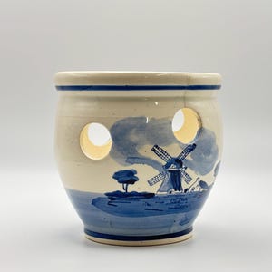 May include: A cream-coloured ceramic jar featuring a blue painted windmill scene. The jar has two circular cutouts and a blue rim. The windmill scene depicts a windmill, trees, and water, all in shades of blue.