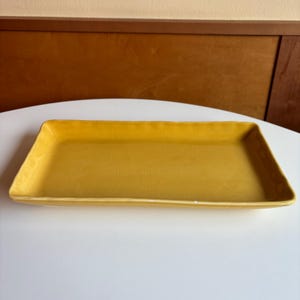 May include: A rectangular ceramic tray in a vibrant mustard yellow color. The surface features a subtle, fine crackle glaze pattern throughout the interior, with slightly raised edges and a smooth, glossy finish on the flat base and sides.