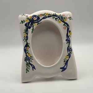 Puede incluir: Un marco de fotos de cerámica blanca con una abertura ovalada. El marco está decorado con una cinta azul y un diseño floral en amarillo, rosa y verde. El marco tiene una forma decorativa.