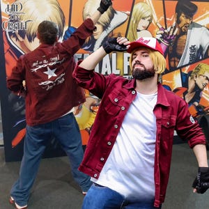 Op de afbeelding: Twee mensen poseren in bordeauxrode spijkerjacks en blauwe jeans voor een kleurrijke muurschildering. Eén jas heeft een witte ster en tekst op de rug. De andere persoon draagt een rood-witte baseballpet en zwarte handschoenen.