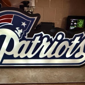May include: A wooden sign featuring the New England Patriots logo and team name in white, blue, and red. The sign is shaped to match the team's logo and is displayed on a countertop.