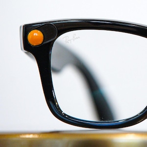May include: Black-framed eyeglasses with clear lenses. The frames have a retro design with a thick, rounded shape. Orange and purple circular accents are on the outer corners of the frames. The Ray-Ban logo is visible on the lens.