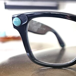 May include: Black-framed eyeglasses with a retro design. The frames have a rounded shape and are accented with small, turquoise-colored embellishments on each corner. The lenses are clear, and the arms are dark.