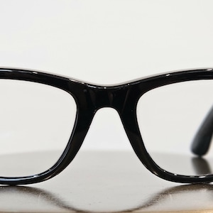 May include: Black-framed eyeglasses with a classic design. The frames have a glossy finish and feature a small, round, orange-colored embellishment on the left side and a green one on the right side. The lenses are clear.