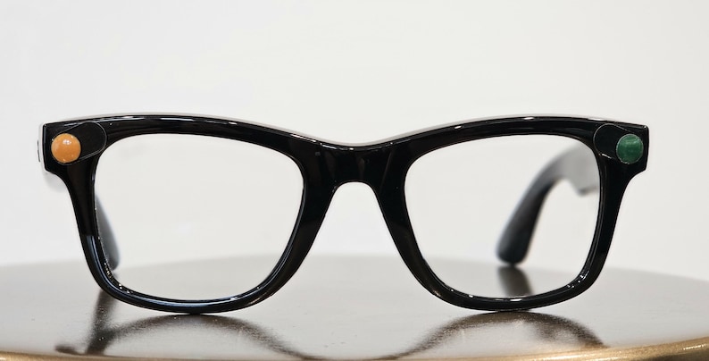 May include: Black-framed eyeglasses with a classic design. The frames have a glossy finish and feature a small, round, orange-colored embellishment on the left side and a green one on the right side. The lenses are clear.