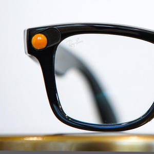 May include: Black eyeglasses with a glossy finish and a prominent orange circular accent on the upper corner of the frame. The clear lens features the Ray-Ban logo printed in white script on the upper right side of the lens surface.