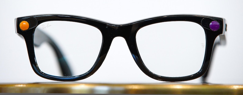 May include: Black-framed eyeglasses with clear lenses. The frames have a retro design with a thick, rounded shape. Orange and purple circular accents are on the outer corners of the frames. The Ray-Ban logo is visible on the lens.