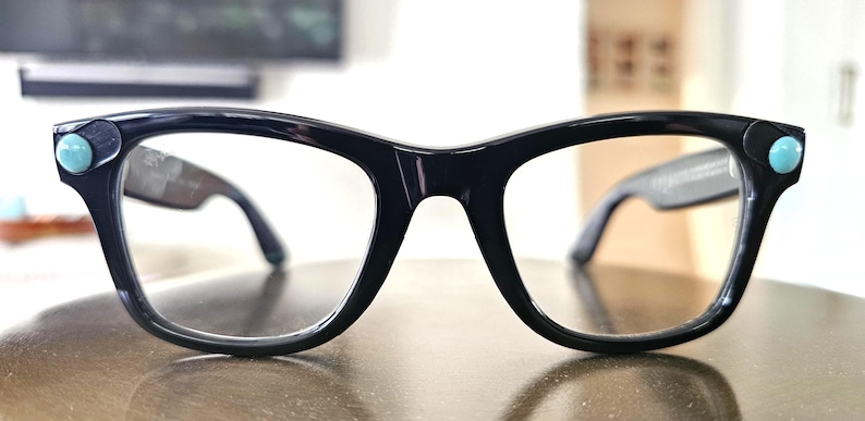 May include: Black-framed eyeglasses with a retro design. The frames have a rounded shape and are accented with small, turquoise-colored embellishments on each corner. The lenses are clear, and the arms are dark.