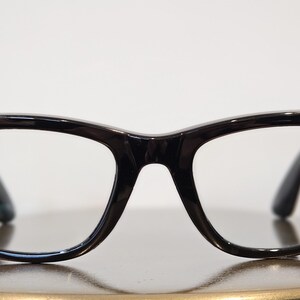 May include: Black-framed eyeglasses with clear lenses. The frames have a retro design with a rounded shape. A blue dot is on the left side of the frame, and a red dot is on the right side. The glasses are on a reflective surface.