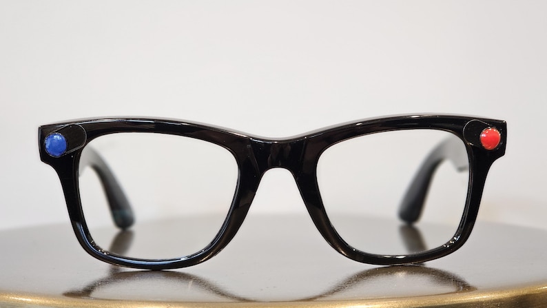 May include: Black-framed eyeglasses with clear lenses. The frames have a retro design with a rounded shape. A blue dot is on the left side of the frame, and a red dot is on the right side. The glasses are on a reflective surface.