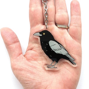 May include: A black and grey crow keyring with a silver chain and clasp. The crow is depicted in a cartoon style with a yellow eye.
