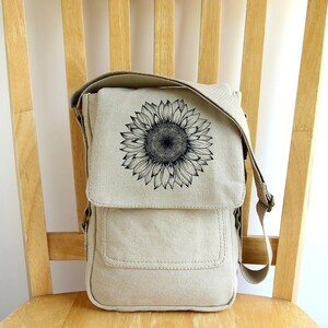 May include: A beige canvas messenger bag with a black sunflower graphic. The bag has a flap closure and an adjustable shoulder strap.