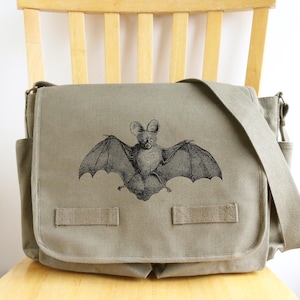 May include: A gray messenger bag with a flap closure and adjustable shoulder strap. The bag features a black and white illustration of a bat with outstretched wings on the front. The bag has two side pockets and is sitting on a wooden chair.