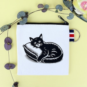 Puede incluir: Un monedero de lona blanca con cremallera negra y una ilustración en blanco y negro de un gato durmiendo sobre una almohada. El monedero tiene un pequeño lazo con rayas rojas, blancas y azules.