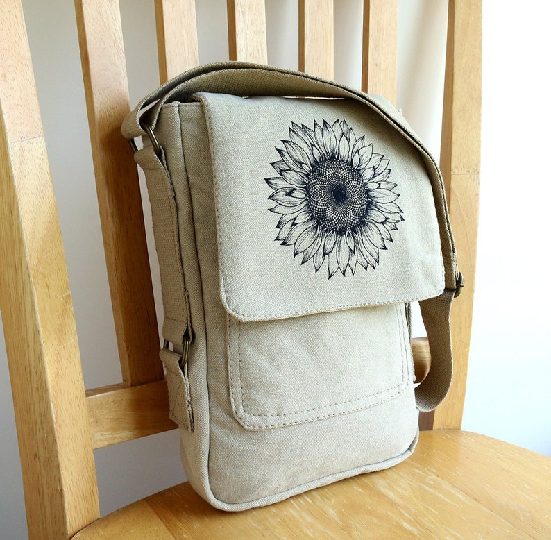 May include: A beige canvas messenger bag with a black sunflower design. The bag has a flap closure and an adjustable shoulder strap.