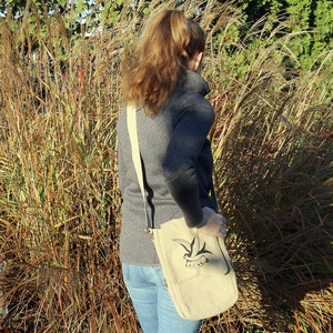 May include: A woman wearing a gray sweater and blue jeans is carrying a beige canvas shoulder bag with a black and white bird design and the word "Sail" printed on it.