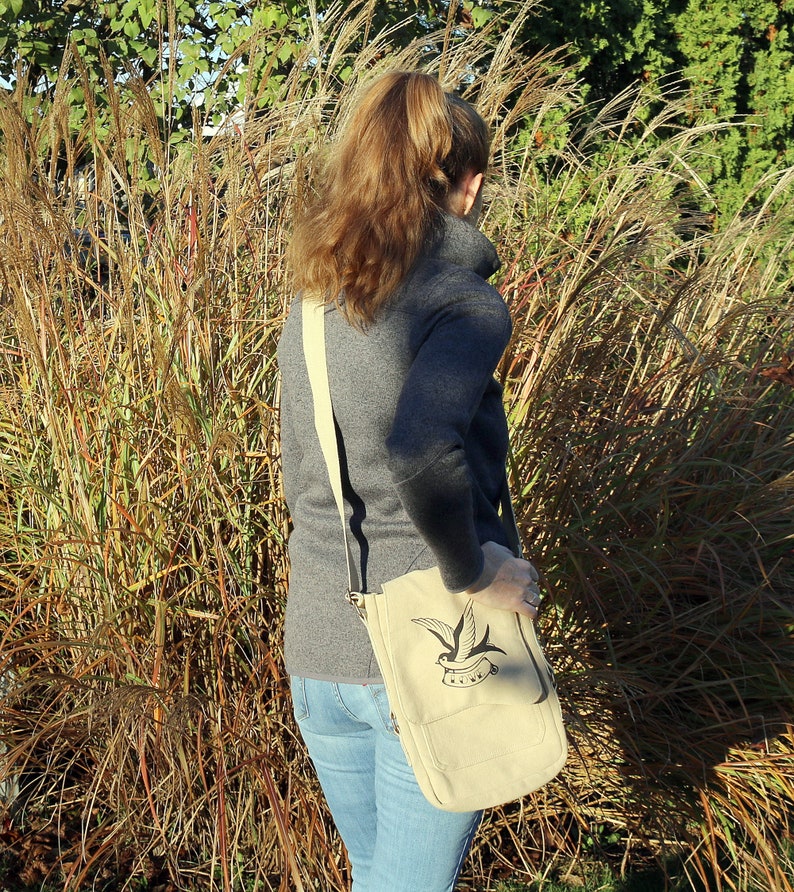 May include: A woman wearing a gray sweater and blue jeans is carrying a beige canvas shoulder bag with a black and white bird design and the word "Sail" printed on it.