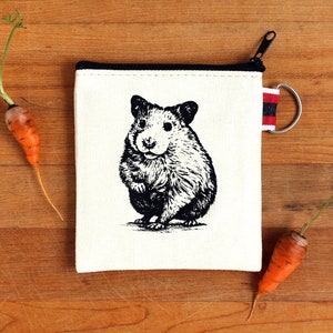 May include: A white canvas zipper pouch with a black zipper and a black and white illustration of a hamster on the front. The pouch has a red and black striped zipper pull.