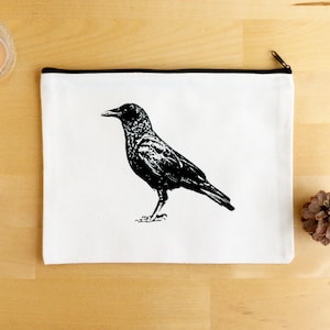 May include: White canvas zipper pouch featuring a black illustration of a crow. The pouch has a black zipper and trim. A lit candle in a glass votive and a pine cone are also visible.