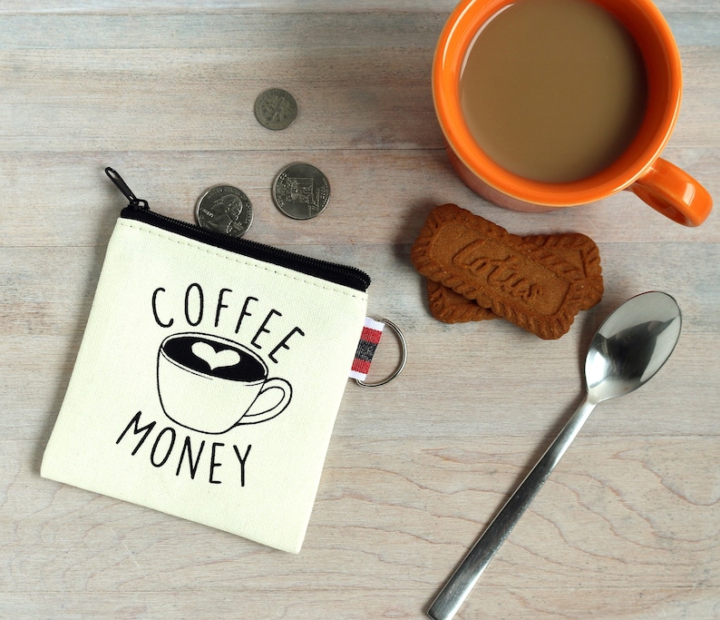 May include: A cream-colored coin purse with the words "COFFEE MONEY" and a coffee cup design. The purse has a black zipper and a small keychain loop. Beside the purse are coins, a mug of coffee, a spoon, and two cookies.