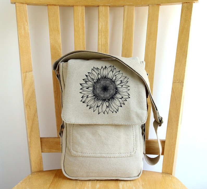 May include: A beige canvas messenger bag with a black sunflower graphic. The bag has a flap closure and an adjustable shoulder strap.