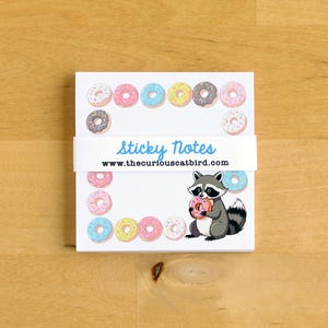 Può includere: Un blocco di note adesive con una fascia bianca con la scritta "sticky notes" e "www.thecuriouscatbird.com". Le note hanno un bordo di ciambelle colorate e un procione dei cartoni animati che tiene in mano una ciambella.