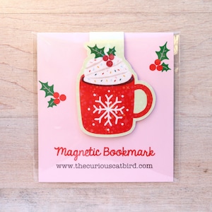 Op de afbeelding: Magnetische boekenlegger met een rood mokontwerp, een sneeuwvlok en slagroomtopping, versierd met hulst en bessen. De boekenlegger staat op een roze achtergrond met de tekst "Magnetic Bookmark" en een websiteadres.