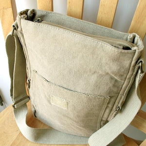 May include: A beige canvas messenger bag with a zippered top and a front pocket. The bag has a long, adjustable shoulder strap and is sitting on a wooden chair.