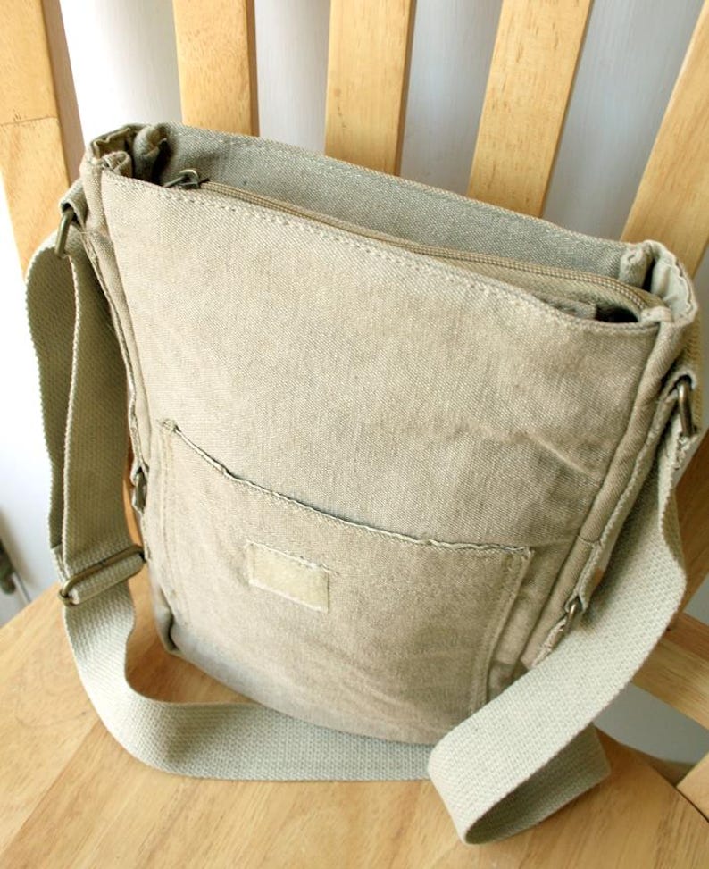 May include: A beige canvas messenger bag with a zippered top and a front pocket. The bag has a long, adjustable shoulder strap and is sitting on a wooden chair.