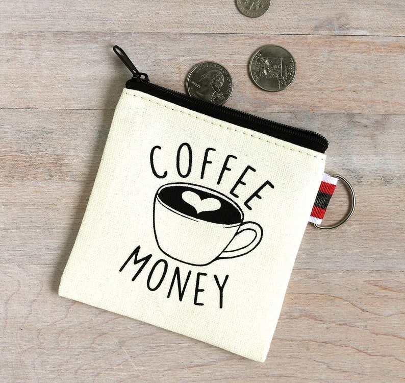 May include: Cream-colored canvas coin purse with a black zipper and a small red, black, and white striped tab. The purse features a black and white graphic of a coffee cup with a heart-shaped design and the words "COFFEE MONEY."