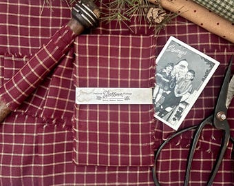 May include: A stack of burgundy fabric with a tan grid pattern, wrapped in a paper band labelled Prairie Blossom Vintage. The fabric is accompanied by a vintage black and white photograph of two children with Santa, metal scissors, and a wooden tool.