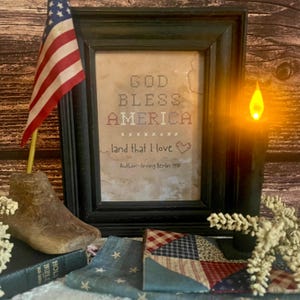 May include: A framed cross-stitch artwork with the text "GOD BLESS AMERICA" and "land that I love." An American flag, a wooden shoe form, and a candle are also present. The artwork is in a black frame.