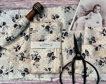 May include: Off-white fabric with a black floral and polka dot pattern, wrapped around a wooden spool. A paper band reads Prairie Blossom Vintage. Beside the fabric are black metal scissors and a vintage photograph of a baby in a white dress.