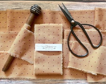 May include: A length of tan fabric with small dark red polka dots, featuring frayed edges. A wooden spool with metal rings sits on the fabric, alongside black metal scissors. A white paper label reads Prairie Blossom Vintage.