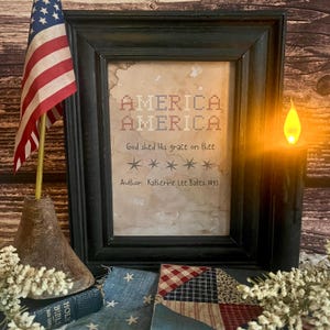 Puede incluir: Una obra de arte enmarcada con las palabras "AMERICA AMERICA" en punto de cruz, y la frase "God shed His grace on thee". Una bandera americana y una vela están al lado del marco. Un pequeño libro y tela están en primer plano.
