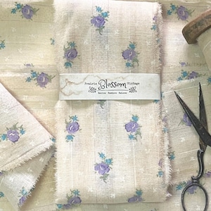 May include: Two pieces of off-white fabric with a small purple and blue floral pattern. The fabric has a slightly textured surface and a frayed edge. A label reads "Prairie Blossom Vintage Revive Restore ReLove".