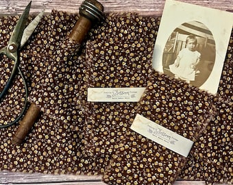 May include: Dark brown fabric with a small yellow and white floral pattern, featuring frayed edges. The fabric is wrapped with white paper bands labelled Prairie Blossom Vintage. Includes metal scissors, a wooden tool, and a vintage photo.